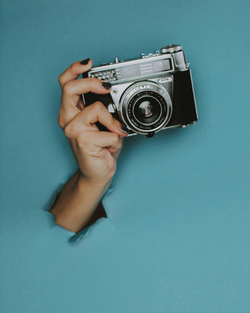 Female hand holding a retro camera through a hole in blue paper.の写真素材