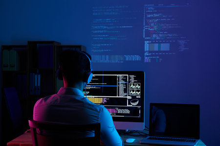 Back view of young man working on computer late at night in office