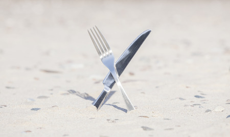 Crossed knife and fork stuck in sand, shallow depth of field.