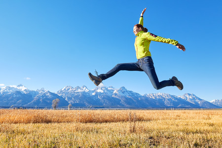 Happy young woman jumping on a meadow, Grand Teton mountain range in distance, Wyoming, USA.の写真素材