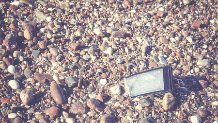 Broken mobile phone with cracked screen on a stony beach, selective focus, color toned picture.の写真素材