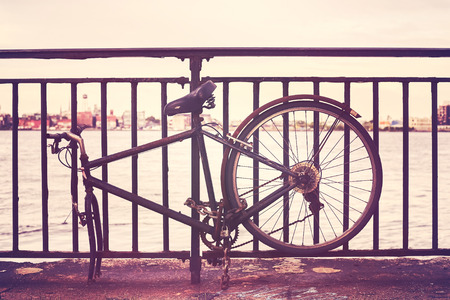 Vintage toned picture of a broken bike fasten to a fence by a river.の写真素材