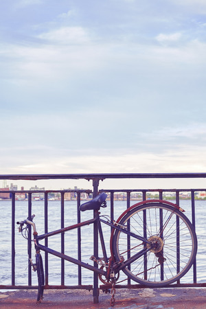 Vintage toned picture of a broken bike fasten to a fence by a river.の写真素材