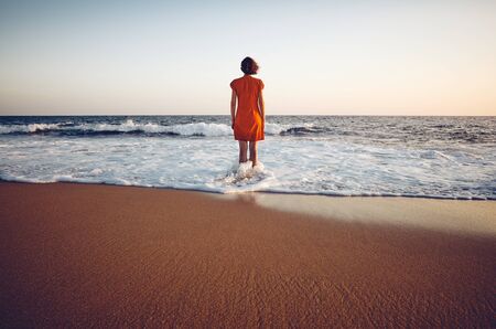 Rear view of a woman in orange dress standing still in the sea at sunset, color toning applied.の写真素材