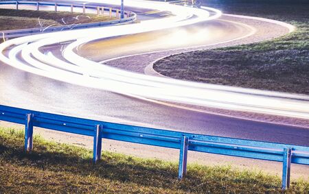Curvy road with car light trails at night, focus on guardrail, color toning applied.の写真素材