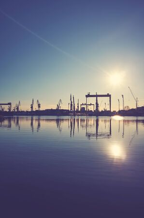 Shipyard cranes silhouettes reflected in water at sunset, color toning applied.の写真素材