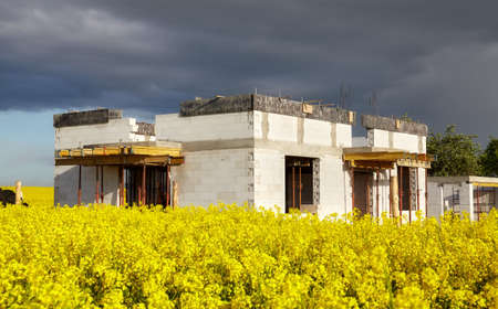 Rural single family house under construction with rapeseed field in foreground and stormy clouds in background.の写真素材