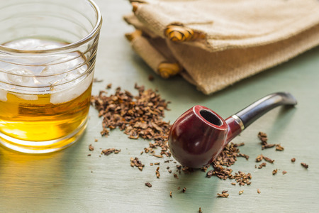 Tobacco pipe on rustic warn green wood surface with spilled natural tobacco and a glass of whisky on...