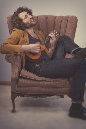 Bearded musician playing acoustic ukulele guitar in vintage chairの写真素材