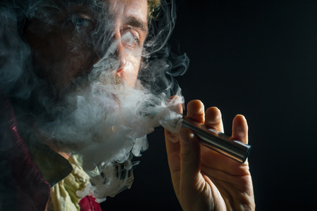 Bearded man smokes a vape e-cigarette on dark backgroundの写真素材
