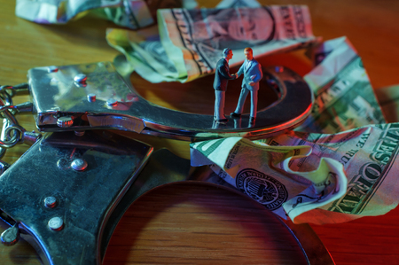 Conceptual miniature criminal businessmen shake hands standing on top of handcuffs with crumpled dollar bills and police lightsの写真素材