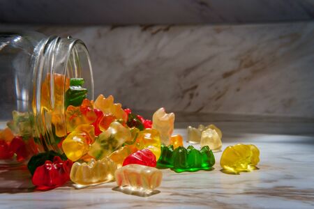 Assorted flavors of gummy bear candies in spilled glass jar on marble kitchen tableの写真素材