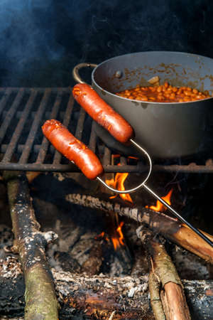 Roasting campfire bratwurst on hot dog fork with cowboy beans slowcooking in backgroundの写真素材