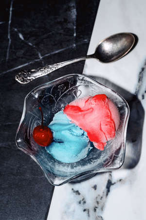 Red and blue scoops of gelato icecream garnished with a maraschino cherry served on black and white marble tableの写真素材