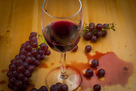 Red wine poured into wine glass and spilled on wooden table with fresh grapes as background design