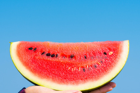A hand holding a slice of watermelon on the beach with blue sea in the background.の写真素材