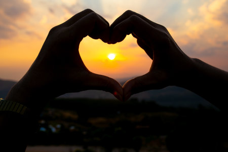 Silhouette of hands forming a heart shape with sunset background.の写真素材