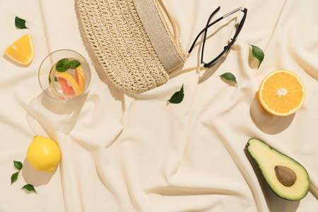 Straw clutch with sunglasses and cocktail glass. Citrus fruits, avocado and green leaves on beige silky fabric.の写真素材