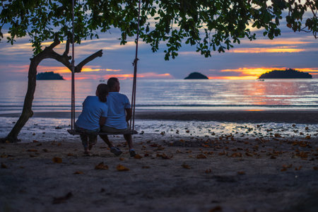 Man and woman sitting on a swing on the beach at sunset.の写真素材