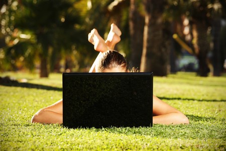 A picture of a barefoot woman on the grass hiding behind her laptop taken during nice sunny dayの写真素材
