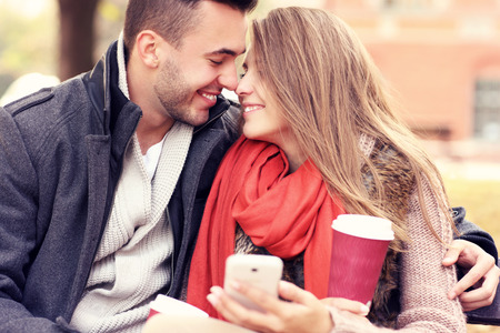 A picture of a young romantic couple kissing on a bench with smartphone in the parkの写真素材