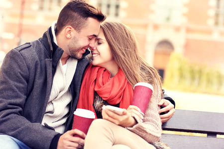 A picture of a young romantic couple kissing on a bench with smartphone in the parkの写真素材