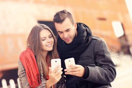 A picture of a young couple sitting on a bench and using smartphones on an autumn dayの写真素材