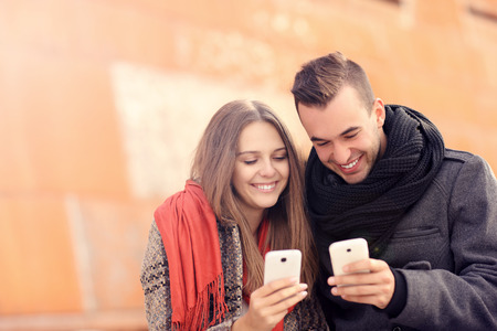 A picture of a young couple sitting on a bench and using smartphones on an autumn dayの写真素材