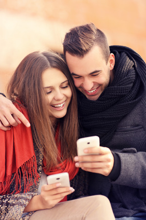 A picture of a young couple sitting on a bench and using smartphones on an autumn dayの写真素材