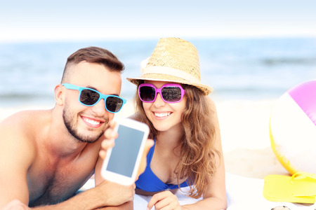 A picture of a happy couple showing smartphone screen at the beachの写真素材
