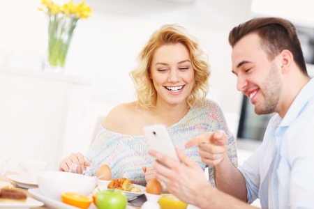 A picture of a young couple eating breakfast in the kitchen and using smart phoneの写真素材