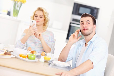 A picture of a young couple eating breakfast in the kitchen and using smart phoneの写真素材