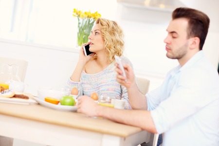 A picture of a young couple eating breakfast in the kitchen and using smart phoneの写真素材