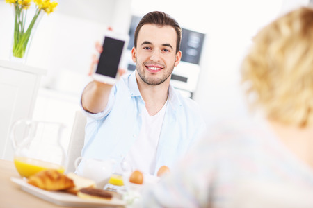 A picture of a happy man showing his smart phone during breakfastの写真素材