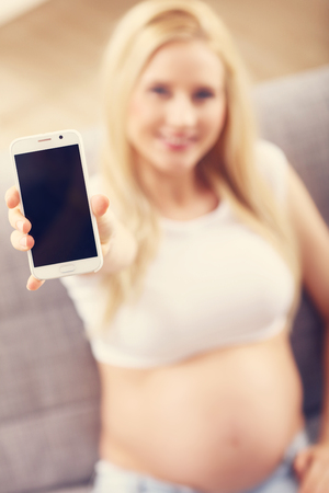 Happy pregnant woman resting on sofa with smartphoneの写真素材