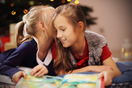 Two cute little sisters reading story book together under Christmas treeの写真素材