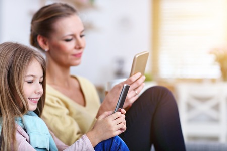 Young mom and her little daughter using smartphone and sitting on sofa at homeの写真素材