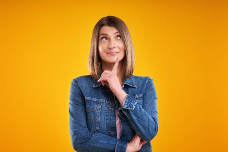 Close up of woman in denim jacket thinking and pointing over yellow backgroundの写真素材