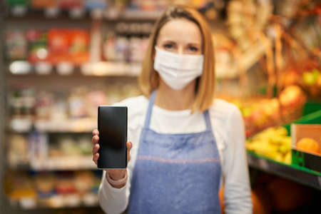 Shop assistant in medical mask holding smartphone against grocery store