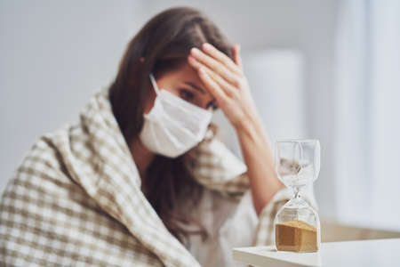 Adult woman in mask feeling sick at home with hourglass