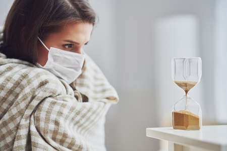 Adult woman in mask feeling sick at home with hourglassの写真素材