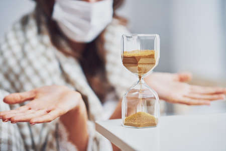 Adult woman in mask feeling sick at home with hourglass
