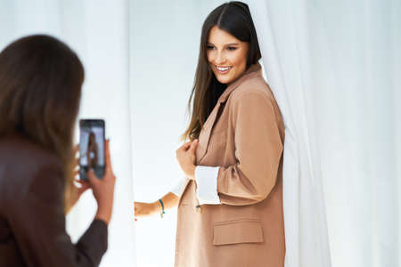 Two girl friends in clothes store in changing room taking selfieの写真素材