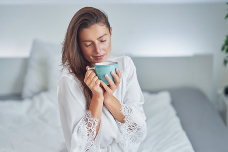 Brunette woman on bad in pajama with mug with coffee or teaの写真素材