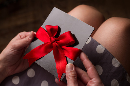 Close up of the hands of a woman with polka dress holding a black card with red ribbon above kneesの写真素材