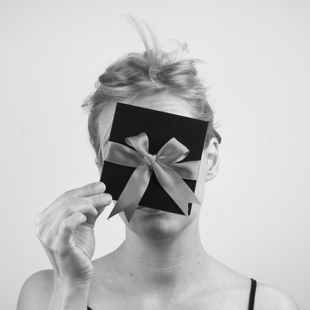 Black and white portrait of a young Caucasian woman holding a black gift card with red ribbon in front of her faceの写真素材