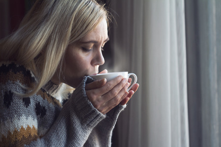 Blonde woman with long hair and warm sweater drinking a cup of hot steamy coffee in the morning by...