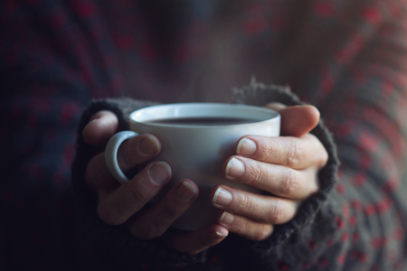 Woman in dark sweater hands holding a white cup of hot coffee with steam coming outの写真素材