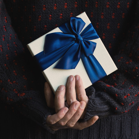 Close up of a man in knitted dark sweater holding a white present gift box with blue ribbon behind his backの写真素材