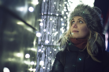 Caucasian blonde woman dressed for winter watching the decorative lled ights on a building wall,...